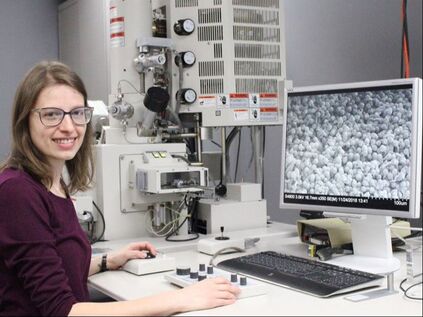 A photo of Wendy Semski, a white, late-20s female with shoulder-length brown hair and glasses. She smiles at the camera, and is seated at a table working with a scanning electron microscope. Her hands are at the controls, and an image of many pollen grains is visible on a computer monitor.